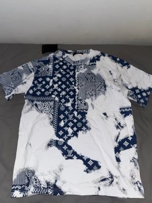 Vit och blå mönstrad t-shirt - Cool t-shirt med unikt blåvitt mönster i bandana-stil och tryck över hela plagget. T-shirten har rund hals och korta ärmar, perfekt för en avslappnad streetwear-look. Materialet känns som bomull och passformen är normal. PASSAR M. FÖRST TILL KVARN