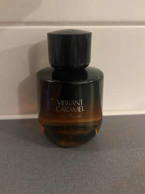 Zara Vibrant Caramel parfym - En elegant parfymflaska från Zara med namnet Vibrant Caramel. Flaskan har en rund form och är gjord av glas i en snygg svart och brun toning. Lock i svart plast. Perfekt för dig som gillar söta och varma dofter.70ml kvar