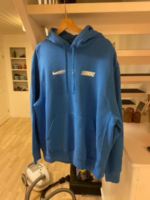 Blå Nike hoodie med tryck - Snygg blå hoodie från Nike med vit logga och coolt tryck framtill. Klassisk känguruficka och justerbar huva med dragsko. Perfekt för dig som vill ha en sportig och avslappnad stil. Tillverkad i mjukt material för extra komfort.
