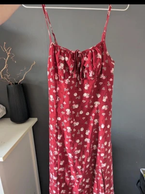 Röd blommig sommarklänning med knyt - Söt röd långklänning med vita blommor och smala axelband. Klänningen har en liten knytning framtill vid bysten och ett avslappnat fall. Perfekt för varma dagar och har en romantisk vibe.