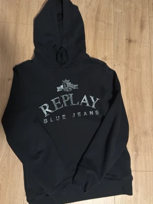 Svart hoodie från Replay - Snygg svart hoodie från Replay med stort tryck 'REPLAY BLUE JEANS' på bröstet och en liten logga ovanför. Klassisk modell med huva och dragsko, samt ribbade muddar vid ärmslut och nederkant. Perfekt för en avslappnad och trendig stil.