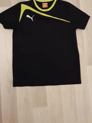 Svart sporttröja från Puma - Snygg svart sporttröja från Puma med korta ärmar och gulgrön detalj över bröstet. Puma-loggan i vitt på framsidan. Tillverkad i ett lätt och funktionellt material som passar perfekt till träning eller sportaktiviteter.