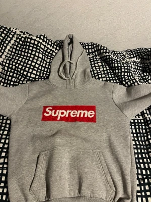 Grå Supreme hoodie med box logo - Säljer en grå hoodie från Supreme med den klassiska röda box loggan broderad på bröstet. Hoodien har huva med dragsko och en stor magficka. Tillverkad i mjukt bomullsmaterial som är perfekt för chill dagar. En riktig streetwear-ikon för dig som gillar hype. Skulle tipa på att den är storlek M till L den är inte XL 