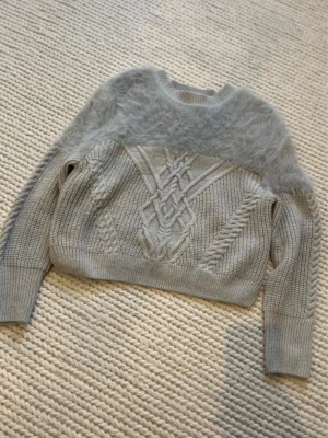 Grå stickad tröja med fluffig detalj - Snygg grå stickad tröja med kabelstickat mönster framtill och breda ribbade muddar. Överdelen har en fluffig, mjuk sektion som ger en unik look. Tröjan har rund hals och långa ärmar. Perfekt för dig som gillar trendiga och mysiga plagg.