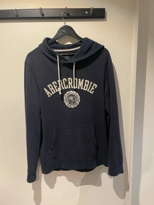  Abercrombie & Fitch - Svart hoodie från Abercrombie & Fitch med vit broderad logga och text på bröstet. Klassisk känguruficka framtill, justerbar huva med vita snören och mjukt bomullsmaterial. Perfekt för en avslappnad och stilren look.