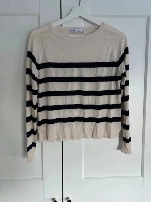 Randig stickad tröja från Zara - Beige tunn stickad tröja från Zara med svarta horisontella ränder. Tröjan har rund halsringning, slits i sidan, långa ärmar och ribbade muddar. Materialet är tunt och mjukt. Sömmen i ena slitsen har gått upp lite (se bild 2)
