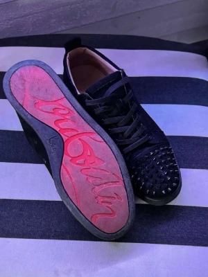 Svarta Louboutin sneakers med nitar - Säljer ett par svarta sneakers från Christian Louboutin med klassisk röd sula och coola svarta nitar på tån. Skorna har snörning, låg profil och är tillverkade i mocka och läder. Perfekta för dig som vill sticka ut med en edgy och lyxig stil.