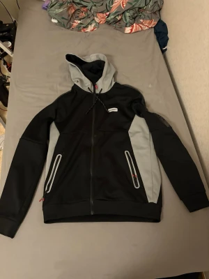 Svart och grå hoodie från McKenzie  - Snygg svart och grå hoodie från McKenzie med dragkedja framtill och två fickor med dragkedja. Huva med dragsko och kontrastfärgade detaljer. Tillverkad i ett mjukt och bekvämt material, perfekt för en avslappnad stil.