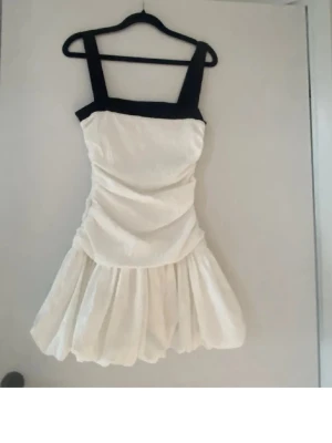 Vit och svart kort klänning - Supersöt kort klänning med breda svarta axelband och överdel, resten av klänningen är vit med lätt rynkad kjol. Perfekt för dig som vill sticka ut med en stilren look. Klänningen har en feminin siluett och fyrkantig ringning.