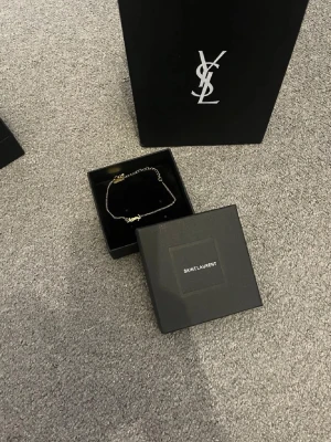 Guldigt halsband från Saint Laurent - Snyggt och stilrent halsband från Saint Laurent i guldtonad metall. Halsbandet har en tunn kedja och ett ikoniskt YSL-hänge framtill. Kommer i original ask och påse, perfekt för dig som gillar exklusiva accessoarer med lyxig känsla.