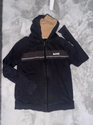 Svart hoodie från BOSS med dragkedja - Snygg svart hoodie från BOSS med dragkedja framtill och beige insida på huvan. Framsidan har ett vitt rutmönster och BOSS-logga tryckt över bröstet. Hoodien har långa ärmar, ribbade muddar och är tillverkad i mjukt bomullsmaterial. Perfekt för en chill och stilren look.