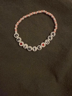 Pärlarmband med text GE INTE UPP - Sött armband med vita och rosa pärlor samt bokstavspärlor som stavar 'GE INTE UPP' med små röda hjärtan. Perfekt accessoar för dig som gillar personliga smycken och vill bära ett peppande budskap. Elastisk tråd för enkel användning.