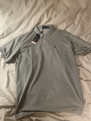 Grå pikétröja från Polo Ralph Lauren - Snygg grå pikétröja från Polo Ralph Lauren med klassisk krage och knappar framtill. Tröjan har korta ärmar och den ikoniska lilla broderade loggan på bröstet. Perfekt för en stilren och avslappnad look. Helt ny! 