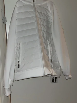 Vit cardiganjacka med huva - Fräsch vit cardiganjacka med quiltat mönster framtill, dragkedja hela vägen och två fickor med dragkedja. Jackan har huva och ribbade muddar vid ärmslut och nederkant. Materialet är en mix av vadderat tyg och mjuk bomullskänsla på ärmarna.