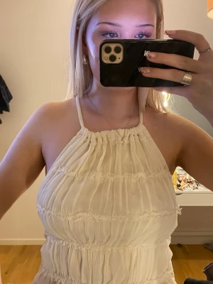 Halterneck öppen rygg - Säljer en ljus, vit Biege  topp med smock och volangdetaljer. Toppen är halterneck med öppen rygg med knyt, den har rynkade volanger och är två lager så den är inte genomskinlig eller obekväm!!