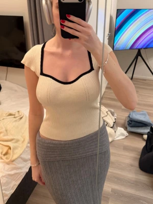 Ribbade topp med svart kant - Snygg ribbad topp i ljus beige med svart kant runt halsringningen. Toppen har korta ärmar och en figurnära passform med en unik fyrkantig urringning. Perfekt för dig som gillar trendiga och stilrena plagg.