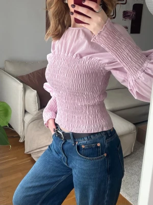 Ljuslila smockad blus med puffärm - Trendig ljuslila blus med smockad kropp och voluminösa puffärmar. Blusen har rund halsringning och är tillverkad i ett lätt tyg som ger en snygg passform. Perfekt för dig som vill ha en feminin och modern look.