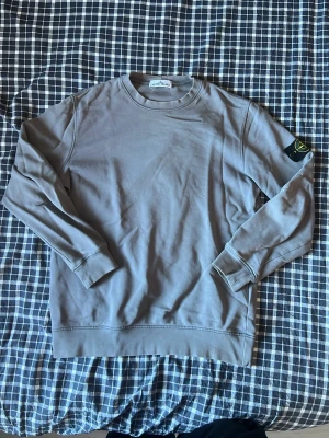 Grå sweatshirt från Stone Island - Säljer en stilren grå sweatshirt från Stone Island med klassisk rund hals och ribbade muddar. Tröjan har den ikoniska Stone Island-loggan på vänster ärm och är tillverkad i mjuk bomull. Perfekt för en avslappnad och trendig look.