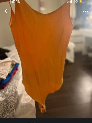 Ribbad orange body med smala axelband - Snygg ribbad body i stark orange färg med smala axelband och rundad urringning både fram och bak. Tillverkad i stretchigt material som sitter tajt mot kroppen. Perfekt att styla med jeans eller kjol för en trendig look.