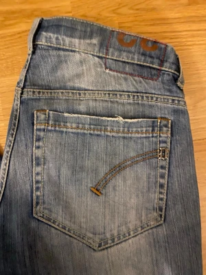 Blå slitna jeans med smal passform - Säljer ett par blå jeans med smal passform och slitna detaljer. Jeansen har klassiska fem fickor, kontrastsömmar och en unik broderad detalj på bakfickan. Materialet är denim i bomull och de har en något tvättad look.