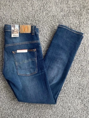 Mörkblå jeans från Nudie Jeans - Snygga mörkblå jeans från Nudie Jeans med klassisk femficksdesign och subtila slitningar framtill. Jeansen har raka ben och orangea sömmar samt en patch med logga bak i midjan. Perfekta för en avslappnad och trendig look.