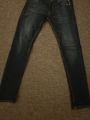 Mörkblå Replay jeans, anbass  - Säljer ett par mörkblå jeans från Replay med smal passform och klassisk femficksdesign. Jeansen har snygga slitningar och kontrastsömmar som ger en cool look. Materialet är denim med stretch för extra komfort. Perfekta för dig som gillar stilrena och trendiga jeans.