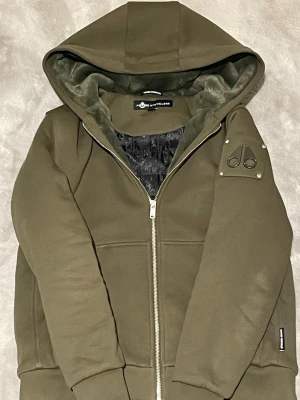 Olivgrön hoodiejacka från Moose Knuckles - Snygg olivgrön hoodiejacka från Moose Knuckles med dragkedja framtill och logotyp på ärmen. Jackan har huva, mjukt foder och ribbade muddar. Perfekt för dig som vill ha en stilren och bekväm jacka med streetkänsla.