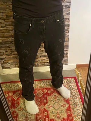 Svarta slitna jeans  - Svarta jeans från SikSilk med distressed detaljer och hål på benen. Klassisk femficksmodell med knappgylf och raka ben. Jeansen har en trendig, avslappnad passform och är tillverkade i bomull med lite stretch för extra komfort.