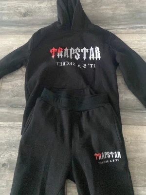 Svart Trapstar set - Svart loungewear set från Trapstar med hoodie och matchande byxor. Hoodien har tryck i rött, vitt och grått med texten 'Trapstar It's a secret' på bröstet. Byxorna har samma tryck på ena benet. Perfekt för chill dagar hemma.