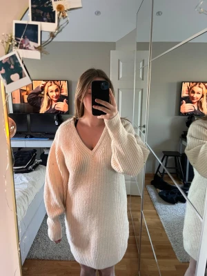 Beige stickad klänning med v-ringning - Mysig stickad klänning i beige med v-ringad hals och långa ärmar. Klänningen har en loose passform och är ribbad, perfekt för att skapa en avslappnad look. Passar dig som gillar enkel och stilren design.