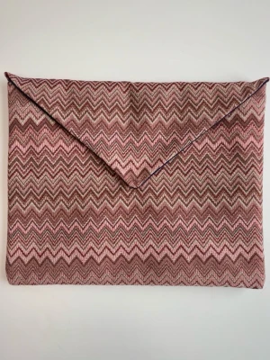 Rosa datorfodral med zigzag-mönster - Snyggt datorfodral i tyg med rosa zigzag-mönster. Väskan har en klassisk kuvertform med spetsigt lock och stängs med en dold knapp. Perfekt accessoar för att lyfta din outfit med lite färg och mönster. De är handgjorda och inuti finns det en ficka samt ett resårband för att organisera pennor (kolla vår tik tok för bilder)