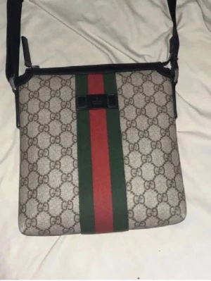 Gucci axelväska  - Säljer en snygg axelväska från Gucci med klassiskt GG-mönster i beige och brunt. Väskan har en bred grön och röd rand i mitten och svart justerbar axelrem. Perfekt för dig som vill ha en trendig och lyxig accessoar till din outfit.