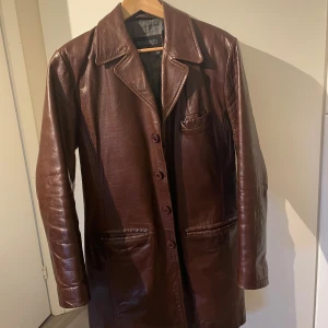 Brun skinnrock från Roco Baroco - Säljer en brun lång äkta skinnrock från Roco Baroco med klassisk krage och knäppning framtill. Jackan har tre ytterfickor och en innerficka med dragkedja. Perfekt för dig som gillar tidlös stil och vill sticka ut. KOM GÄRNA MED EGNA PRISFÖRSLAG!