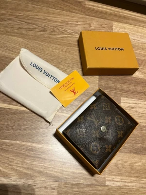 Louis Vuitton monogram plånbok brun - Säljer en brun plånbok från Louis Vuitton med klassiskt monogrammönster i guld och beige. Plånboken är tillverkad i tåligt canvasmaterial och har en knappstängning. Kommer med original ask, dustbag och certifikat. Perfekt för dig som gillar lyxiga accessoarer.