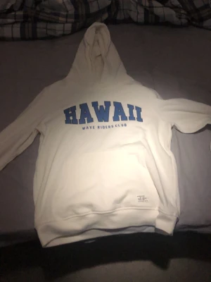 Vit hoodie HAWAII Wave Riders Club - Säljer en vit hoodie med blå text 'HAWAII' och 'Wave Riders Club' på bröstet. Klassisk känguruficka framtill och stor huva. Mjuk bomullskänsla och avslappnad passform, perfekt för chill dagar eller när du vill ha en clean look.