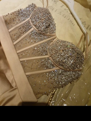 Champagnefärgad långklänning med paljetter - Elegant långklänning i champagnefärg med smala axelband och glittrig överdel täckt av paljetter och pärlor. Klänningen har markerad midja, slits framtill och en mjukt draperad kjol i satinliknande material. Perfekt för bal eller fest. Lite slitningar i släpet, vanligt vid användning