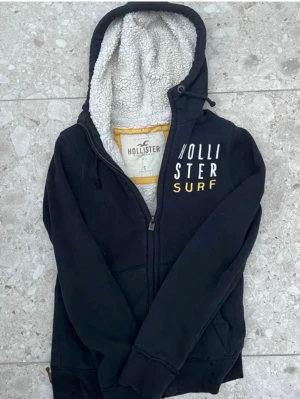 Svart Hollister hoodie med foder - Mysig marinblå hoodie från Hollister med dragkedja och broderad text 'Hollister Surf' i vitt och gult på bröstet. Insidan av huvan är fodrad med vit teddy för extra komfort. Klassisk känguruficka framtill och justerbar huva med snören.