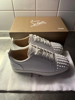 Vita Louboutin sneakers med nitar - Snygga vita sneakers från Christian Louboutin med coola vita nitar på tån. Skorna har en klassisk låg siluett, vita skosnören och är tillverkade i slätt läder. Perfekta för dig som vill sticka ut med en lyxig och edgy stil.