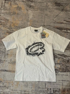 Vit t-shirt från Corteiz - Vit t-shirt från Corteiz med svart grafiskt tryck på bröstet. Klassisk passform med rund hals och korta ärmar. Perfekt för dig som gillar streetwear och vill ha en enkel men snygg look.