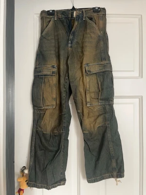 BDG cargobyxor i tvättad denim - Säljer ett par cargobyxor från BDG Urban Outfitters i tvättad blå denim med slitna detaljer och stora fickor på sidorna. Byxorna har en avslappnad passform och cool vintage-look. Perfekta för en streetwear-stil.