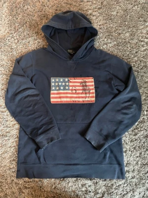 Mörkblå hoodie Ralph Lauren hoodie - Mörkblå hoodie från Polo Ralph Lauren med amerikanska flaggan och en polospelare på bröstet. Klassisk huva och ribbade muddar. Perfekt för en avslappnad och cool stil.