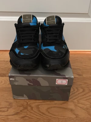 Valentino blå camo sneakers - Snygga sneakers från Valentino med blå och svarta camo-detaljer. Skorna har svarta snören, svart sula och en cool, sportig design. Märkeslogga på plösen och ovandel i textil och läder. Perfekta för dig som vill sticka ut med en unik stil.