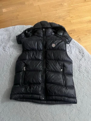 Svart dunväst från Moncler - Snygg svart dunväst från Moncler med klassisk logga på bröstet. Västen har dragkedja framtill, två fickor med dragkedja och en hög krage. Tillverkad i glansigt material och fylld med dun för extra värme. Perfekt för lager-på-lager-stil. Den är inte så sliten