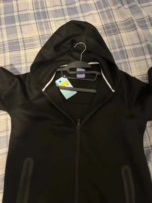 Svart Nike NOCTA hoodie med dragkedja - Svart hoodie från Nike NOCTA-kollektionen med hel dragkedja framtill, två fickor med dragkedja och gul Nike-logga på bröstet. Huva med vit kant och NOCTA-tryck på ärmen. Perfekt för en sportig och stilren look.