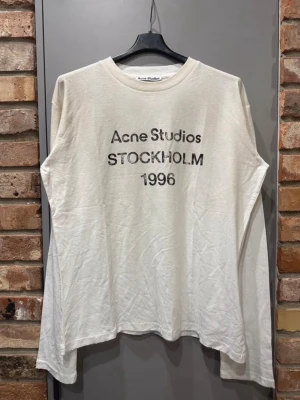Vit långärmad topp Acne Studios - Vit långärmad topp från Acne Studios med tryck 'Acne Studios STOCKHOLM 1996' på bröstet. Klassisk rund halsringning och avslappnad passform. Perfekt till jeans för en clean och trendig look.