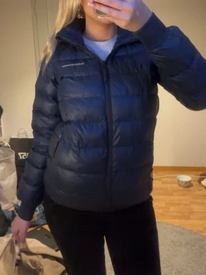 Mörkblå dunjacka från Peak Performance - Snygg mörkblå dunjacka från Peak Performance med huva och dragkedja framtill. Jackan har två sidofickor med dragkedja och quiltad design som ger en puffig look. Perfekt för kalla vinterdagar och riktigt skön att bära.