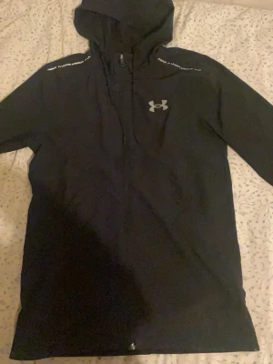 Svart jacka från Under Armour - Svart zip-hoodie från Under Armour med huva och dragsko. Logga i vitt på bröstet och textdetaljer på axlarna. Perfekt för en sportig och avslappnad stil. Mjukt material och hel dragkedja framtill. Inga skador förutom att det finns liten hål i framsidan som knappt syns och lite skada på loggan