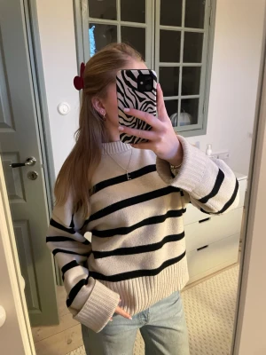 Beige stickad tröja med svarta ränder - En oversized stickad tröja i beige med breda svarta horisontella ränder. Tröjan har ribbstickad hals, muddar och nederkant. Perfekt för att skapa en avslappnad och trendig look. Materialet ser ut att vara mjukt och bekvämt.