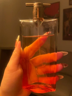 Lancome Idole Now 50ml - Snygg parfymflaska i genomskinligt glas med en varm orange ton. Flaskan har en unik, rektangulär form och ett metallfärgat spraymunstycke. Perfekt för dig som gillar stilrena och moderna detaljer på parfymhyllan.
