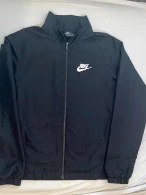 Svart vindjacka från Nike - Snygg svart vindjacka från Nike med dragkedja framtill och vit Nike-logga på bröstet. Jackan har hög krage och elastiska muddar vid ärmsluten. Tillverkad i lätt och tåligt material som passar perfekt för blåsiga dagar.